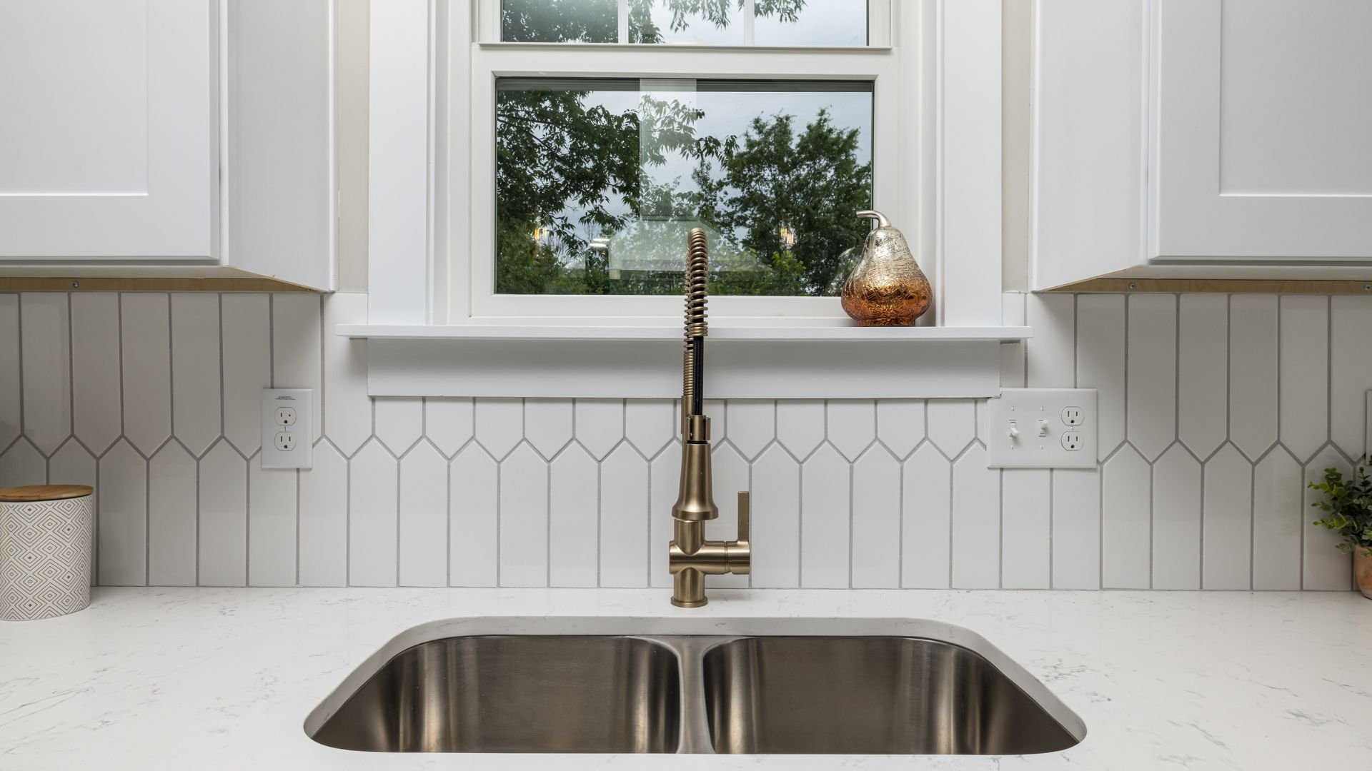 Modern kitchen sink with bronze faucet, white tile backsplash, and window view