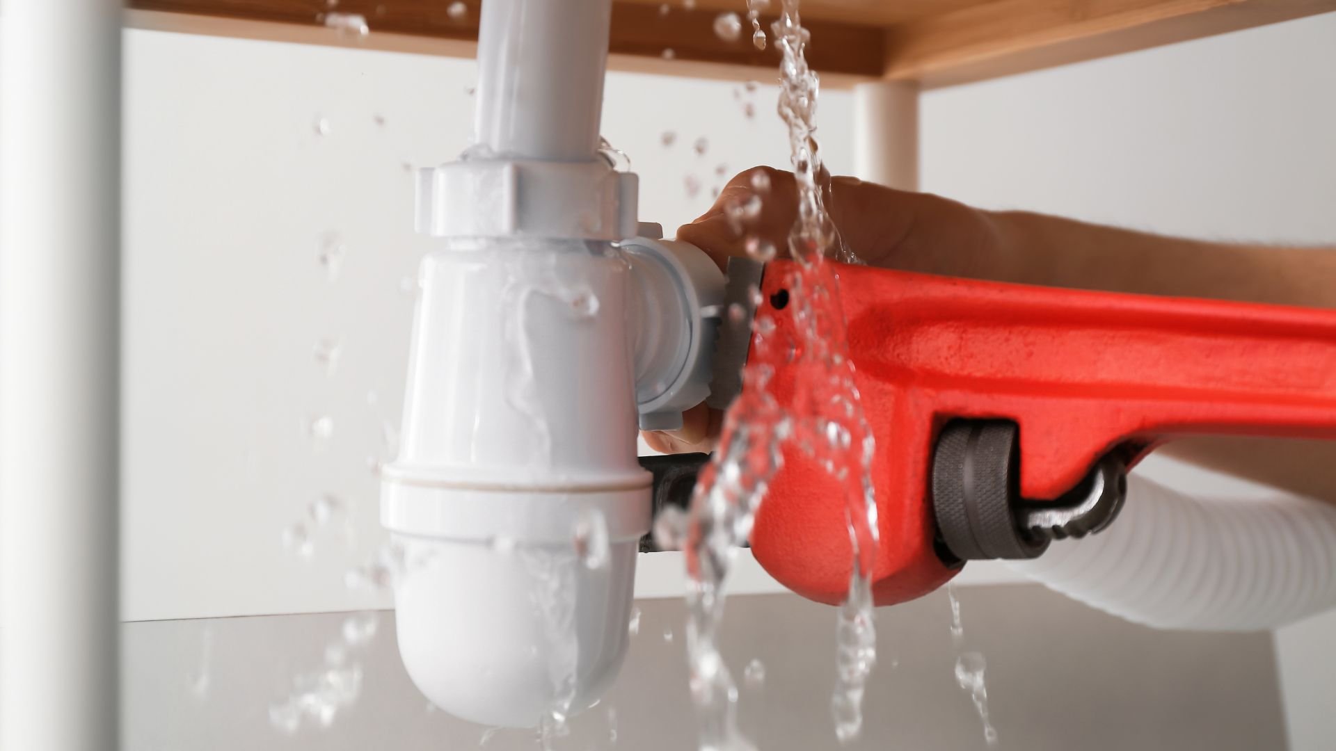 Red pipe wrench fixing leaking white plumbing pipe with water spraying