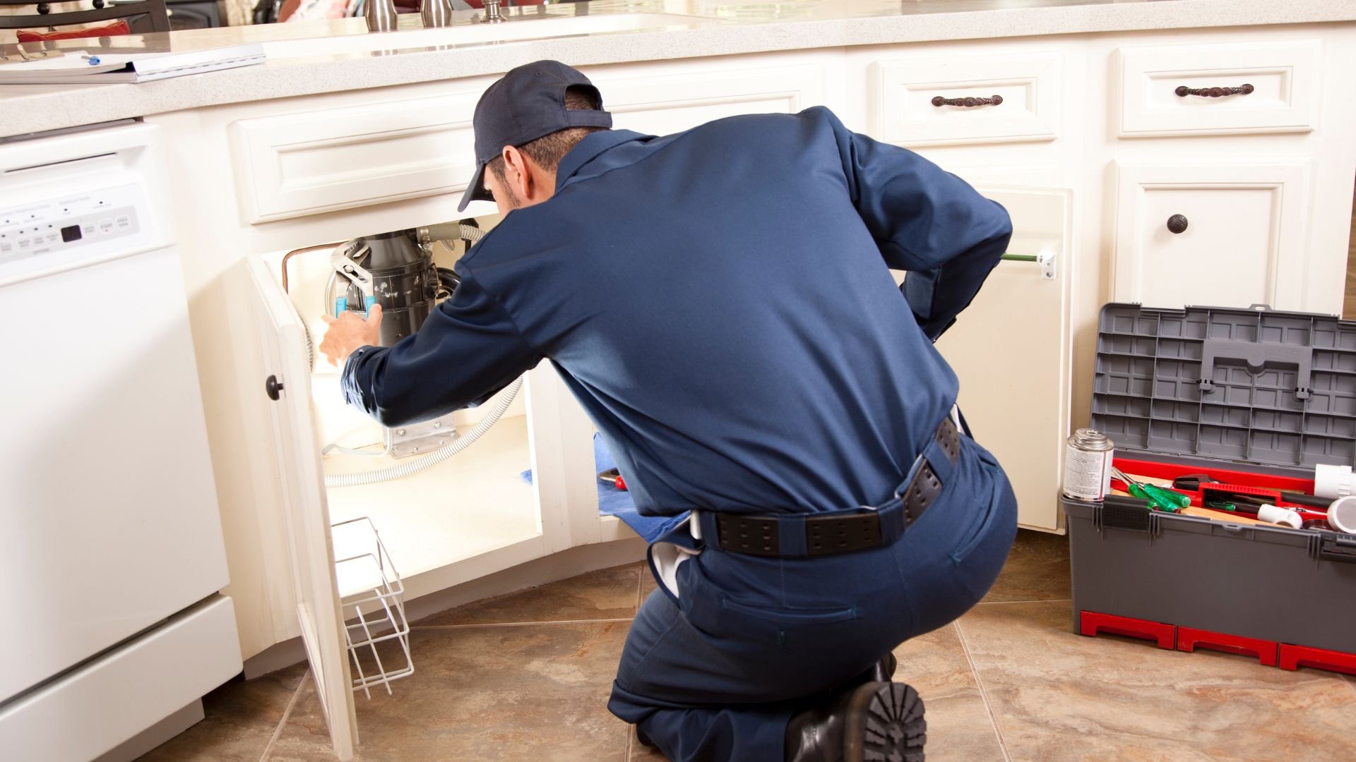 Technician in blue uniform repairs under-sink plumbing in kitchen